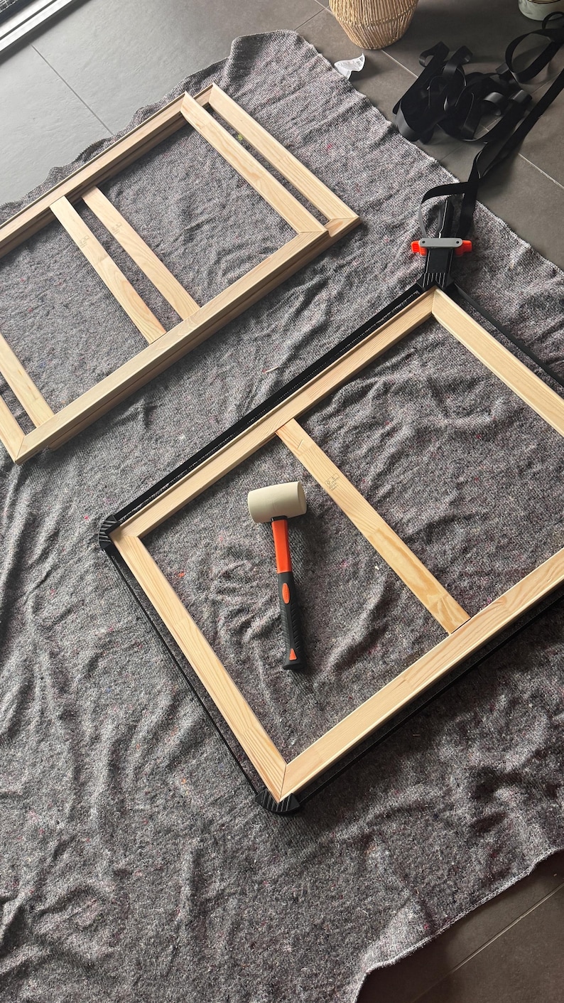 May include: Two rectangular light-colored wooden frames with a central support bar, a black nylon strap clamp secured around one frame, and a rubber mallet with an orange handle and white head resting in the center of the clamped frame.