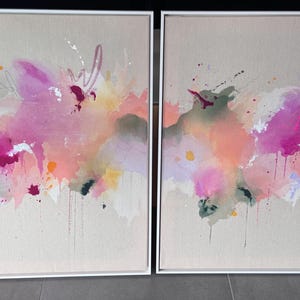 May include: A pair of rectangular framed wall art pieces featuring abstract paint splatters and washes in shades of bright pink, soft peach, muted green, and white. The canvases are set within thin white frames, displaying fluid, layered brushwork.