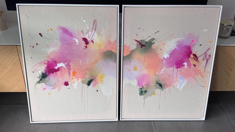 May include: A pair of rectangular framed wall art pieces featuring abstract paint splatters and washes in shades of bright pink, soft peach, muted green, and white. The canvases are set within thin white frames, displaying fluid, layered brushwork.