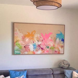 May include: A rectangular canvas painting featuring abstract splashes of neon pink, bright orange, turquoise, and lime green paint on a tan background, framed in light wood. Above it hangs a tiered, circular pendant light with woven cane detailing.