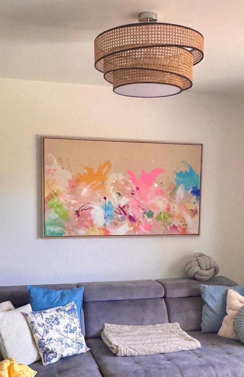 May include: A rectangular canvas painting featuring abstract splashes of neon pink, bright orange, turquoise, and lime green paint on a tan background, framed in light wood. Above it hangs a tiered, circular pendant light with woven cane detailing.