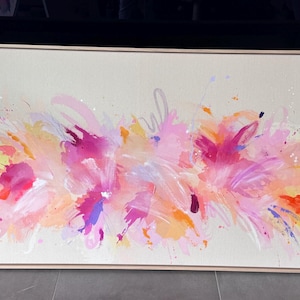 May include: A rectangular canvas painting in a light wood frame featuring an abstract composition of vibrant pink, orange, yellow, and purple paint strokes and splatters on a neutral beige background, creating a dynamic and layered visual effect.