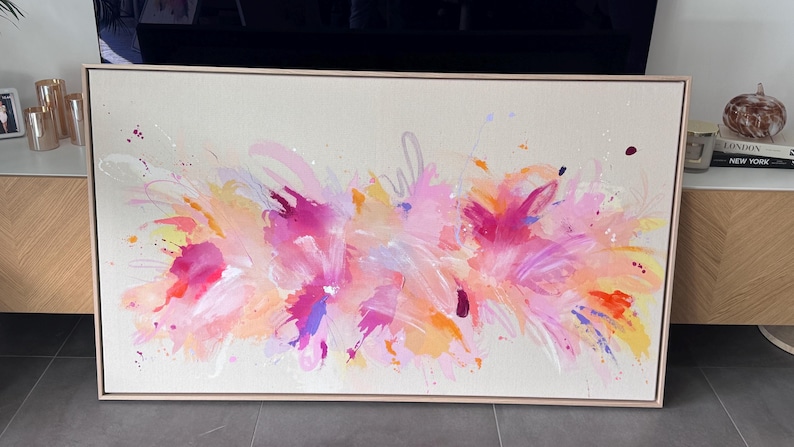 May include: A rectangular canvas painting in a light wood frame featuring an abstract composition of vibrant pink, orange, yellow, and purple paint strokes and splatters on a neutral beige background, creating a dynamic and layered visual effect.