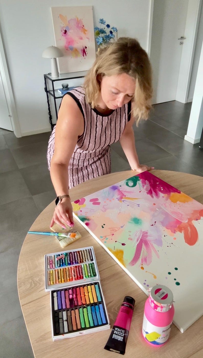 May include: A sleeveless pink dress with black vertical stripes, a rectangular canvas featuring abstract pink, orange, and green paint splatters, a two-tier box of colorful oil pastels, a tube of magenta acrylic paint, and a pink paint bottle.