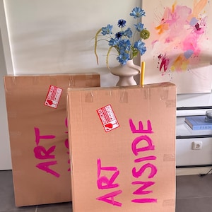 May include: Two large rectangular cardboard shipping boxes with the words ART INSIDE written in bright pink paint. Each box features a red and white fragile sticker with the German text ZERBRECHLICH. The boxes are sealed with clear tape.