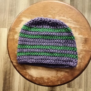 May include: A handmade crochet beanie in shades of purple and green. The hat features horizontal stripes of alternating colors. The beanie is displayed on a wooden stool. This item is perfect for cold weather.