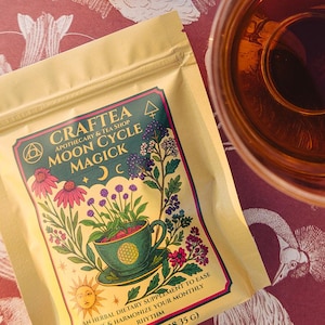 Könnte beinhalten: Ein Stillleben mit einer Teetasse, einer Packung "Craftea Moon Cycle Magick" Tee, rosa Blüten und Kristallen auf einer gemusterten Oberfläche. Die Teepackung hat Blumenillustrationen und Text. Der Tee hat eine tiefe Bernsteinfarbe.