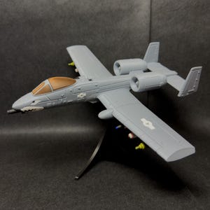 May include: A gray model airplane with a brown cockpit and white shark teeth design on the nose. The model has two engines, wings, and a tail. It is mounted on a black stand and has small colorful details.