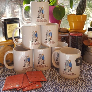 May include: Six white ceramic mugs with a graphic of two people sitting at a sink with the text "Planes, Trains and Pottery Wheels". The mugs are stacked in a pyramid shape on a blue and white patterned tablecloth.