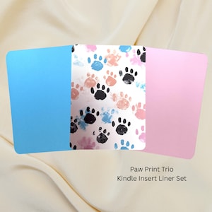 May include: A set of three Kindle insert liners. The center liner features a white background with paw prints in black, pink, and blue. The other two liners are solid, one light blue and one pink. The text reads "Paw Print Trio Kindle Insert Liner Set."