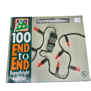 Pode incluir: Uma caixa de 100 luzes de corda vermelhas com fio verde, rotulada 100 END to END SuperBright LightSet. A embalagem apresenta um design verde e laranja com um gráfico de árvore de Natal e uma marca de certificação UL na frente.