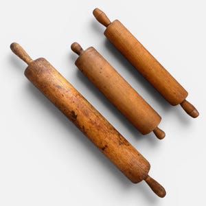 May include: Three wooden rolling pins of different sizes, set diagonally on a white background. The rolling pins are made of light brown wood with visible grain and dark patches, indicating age and use. The handles are rounded and attached to the main body.