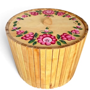 May include: A wooden storage container with a lid. The lid and the top of the container are decorated with pink flowers and green leaves. The container is made of vertical wooden slats.