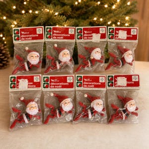 May include: Eight individual Woolcrest Christmas ornaments packaged in clear plastic bags. Each ornament features a small Santa Claus figure with a red hat, white beard, and red suit. The header cards are red and white with the text décorations de noël.