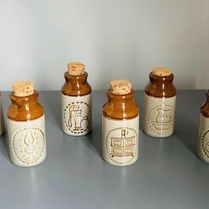 Peut inclure: Un ensemble de six pots cylindriques en céramique avec des cols émaillés marron et des corps beige clair, chacun surmonté d'un bouchon en liège naturel. Chaque pot présente une illustration en relief d'un objet ménager ancien, dont une lampe.