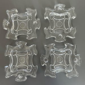May include: Four clear glass ashtrays with a textured surface and a unique, puzzle-piece-like shape. Each ashtray features a central circular indentation and four curved extensions. The ashtrays are arranged on a grey surface.