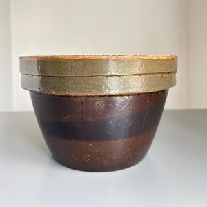 May include: A round ceramic bowl featuring a wide, textured olive-green rim and a body decorated with alternating horizontal bands of dark brown and medium brown. The bowl has a smooth, glazed finish and a slightly tapered shape toward the base.