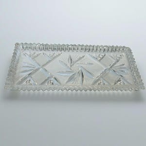 May include: A rectangular, clear glass tray with a cut crystal design. The tray features a diamond and leaf pattern in the centre and a sawtooth edge. This decorative tray is suitable for serving or display.