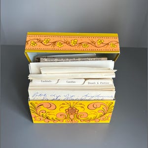 May include: A yellow metal recipe box with an orange floral border on the lid and a decorative orange scroll pattern on the front. The box is filled with white index cards, including dividers labelled Cocktails, Candies, and Breads & Pastries.