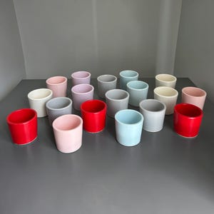 May include: A collection of seventeen small, cylindrical ceramic cups arranged in a group. The cups feature a smooth, matte finish and come in a variety of solid colours, including shades of red, light blue, soft pink, cream, and light grey.
