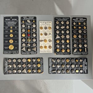 May include: Seven vintage display cards from Defiance Button Machine Co. featuring an assortment of metal buttons in various shapes, sizes, and finishes, including gold, silver, and coloured accents, with printed text labels and model numbers.