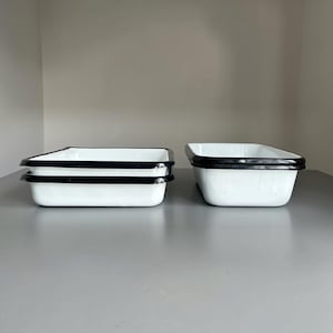 May include: Two rectangular white enamel baking pans with black rims. One pan is stacked on top of another on the left, while a single pan sits on the right. The pans have smooth, glossy surfaces and deep sides, featuring a clean, functional design.