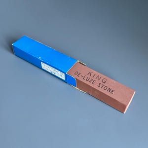 May include: A rectangular brown sharpening stone labelled KING DE-LUXE STONE, partially extended from its bright blue cardboard sleeve. The stone has a smooth, matte surface and is positioned diagonally against a solid grey background.
