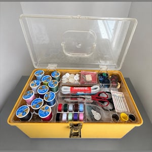 May include: A yellow plastic sewing organiser box with a clear hinged lid containing various spools of thread in multiple colours, white buttons, a red package of needles, elastic, red-handled scissors, and two yellow thimbles inside compartments.