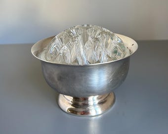 May include: A silver-toned metal bowl featuring a rounded base and a wide rim, containing a clear glass flower frog insert. The glass insert is dome-shaped with multiple circular openings designed to hold individual flower stems upright.