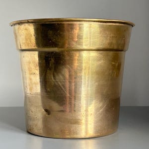 May include: A polished brass planter with a wide, circular rim and a tapered cylindrical body. The metal surface displays a warm, golden-yellow hue with subtle variations in tone and light reflections across its smooth, metallic exterior.