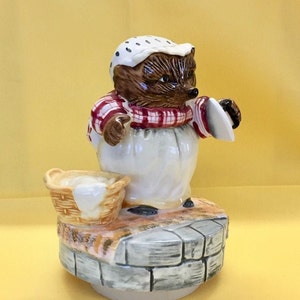 May include: A ceramic figurine of a hedgehog dressed as a baker, wearing a white hat with black dots and a red and white checkered shirt. The hedgehog is standing on a stone well, holding a spatula and a basket.