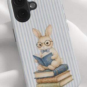 May include: A phone case featuring a light blue and white vertical striped background. The design displays a tan rabbit wearing round glasses and a blue bow tie, sitting on a stack of three books while reading a blue book held in its paws.
