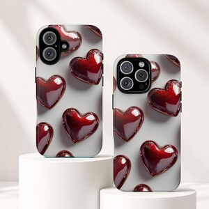 May include: Two smartphone cases featuring a repeating pattern of glossy, deep red, three-dimensional heart shapes against a light gray background. Each case has a prominent camera cutout area at the top left, showcasing the device's lens configuration.