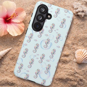 May include: A light blue phone case featuring a pattern of colorful seahorses, bubbles, and small fish. The case has a triple-lens camera and is surrounded by sand, a pink hibiscus flower, and seashells.