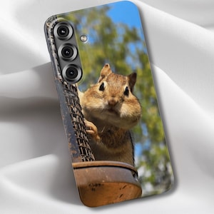 May include: A phone case featuring a close-up image of a chipmunk with puffed cheeks, clinging to a metal bird feeder. The background shows a blurred view of green trees and a blue sky.