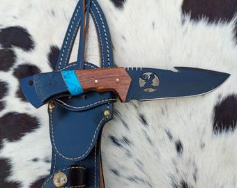 May include: A black folding knife with a wooden handle and a turquoise accent, displayed in a black leather sheath. The knife blade features a deer head emblem. The sheath is stitched with white thread and has a snap closure.