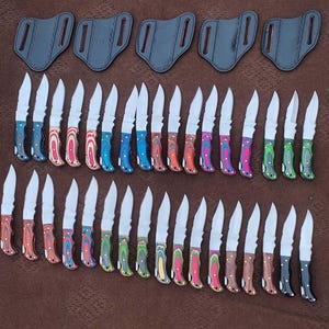 Op de afbeelding: Een assortiment kleine messen met kleurrijke houten handvatten en zilveren bladen, samen met bijpassende leren scheden. De handvatten hebben een verscheidenheid aan kleuren, waaronder blauw, rood, groen en bruin.