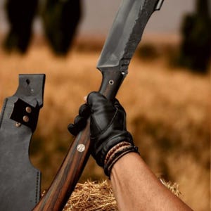Könnte beinhalten: Ein großes Messer mit gebogener Klinge und dunkler, strukturierter Oberfläche. Der Griff besteht aus dunklem Holz mit geschwungenem Design. Das Messer wird in einem schwarzen Lederhandschuh gehalten, mit einer sichtbaren Lederscheide. Der Hintergrund ist eine verschwommene Außenszene.