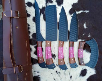 Pode incluir: Um conjunto de seis facas de cozinha com lâminas escuras, cabos de madeira e detalhes rosa, apresentado com um estojo de couro castanho. As lâminas têm um acabamento texturizado. O estojo tem um fecho de fivela.