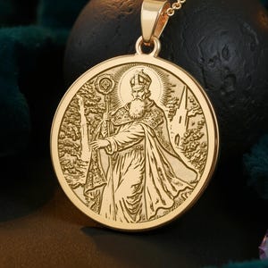 May include: Gold-colored pendant necklace featuring a detailed engraving of a religious figure. The round pendant hangs from a delicate chain. The design includes a figure with a staff, set against a backdrop of trees and a building.