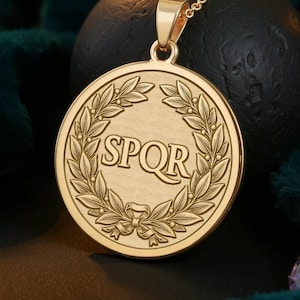Könnte beinhalten: Eine goldfarbene Halskette mit Anhänger, die ein rundes Medaillon zeigt. Das Medaillon trägt die Buchstaben "SPQR" in einem Lorbeerkranz-Design. Die Halskette wird auf dunklem, grünem Samt präsentiert.