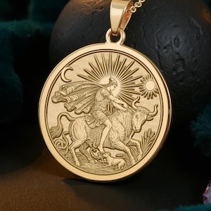 May include: Gold-colored pendant necklace featuring a detailed engraving of a figure riding a bull, with sun and moon symbols. The pendant hangs from a delicate chain. The design is set against a dark background.