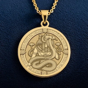 Może przedstawiać: Złoty naszyjnik z wisiorkiem z egipskim wzorem. Okrągła zawieszka przedstawia szczegółowy relief kobry i hieroglifów. Naszyjnik zawieszony jest na delikatnym łańcuszku, na ciemnoniebieskim tle.