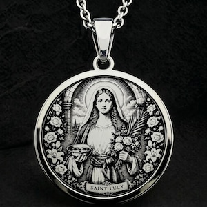 Op de afbeelding: Een ronde zilverkleurige hanger met een gravure van de heilige Lucia die een kleine schaal met ogen en een palmtak vasthoudt. Het ontwerp is omlijst door een bloemenrand en bevat onderaan de tekst Saint Lucy.