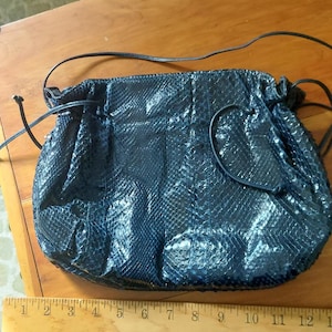 May include: A dark blue, textured handbag with a drawstring closure and a long, thin strap. The bag has a snakeskin pattern and is lying on a wooden surface. The text "Big Bad John" is visible on a ruler.