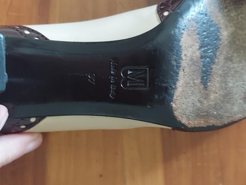 May include: Close-up of a beige and brown women's shoe. The shoe has a black sole with the text "Made in Italy" and the number "37". The shoe has a small heel and a brown leather accent.