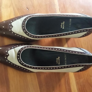 May include: Two-tone brown and cream flats with a wingtip design. The shoes feature decorative perforations and a rounded toe. The interior is black, and the soles are dark brown. Made in Italy is printed on the insole.