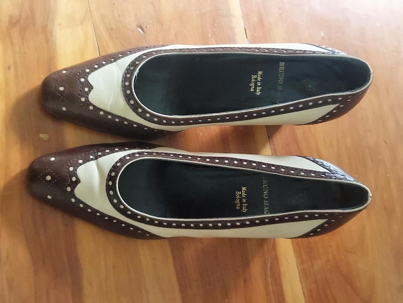 May include: Two-tone brown and cream flats with a wingtip design. The shoes feature decorative perforations and a rounded toe. The interior is black, and the soles are dark brown. Made in Italy is printed on the insole.