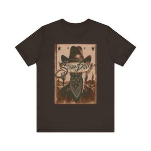 May include: Brown t-shirt featuring a graphic of a cowboy with a bandana covering their face, a cowboy hat, and the text "Straw Billy". The design includes stars and desert cacti in a vintage style.