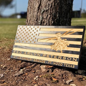 May include: Wooden American flag with engraved stars and stripes, featuring a helicopter and the name "SSG Arlyn Peterson" along with "1st Air Cavalry Brigade - Ft. Hood Texas". The flag is made of wood with a black border.